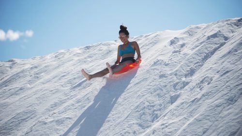 A person in a blue top slides down a snowy slope on a red sled under a clear blue sky.
